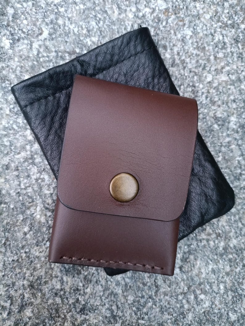 May include: A smooth dark brown leather card holder with a single brass snap button closure, featuring visible stitching along the bottom edge, layered over a black textured leather pouch with a drawstring top closure.