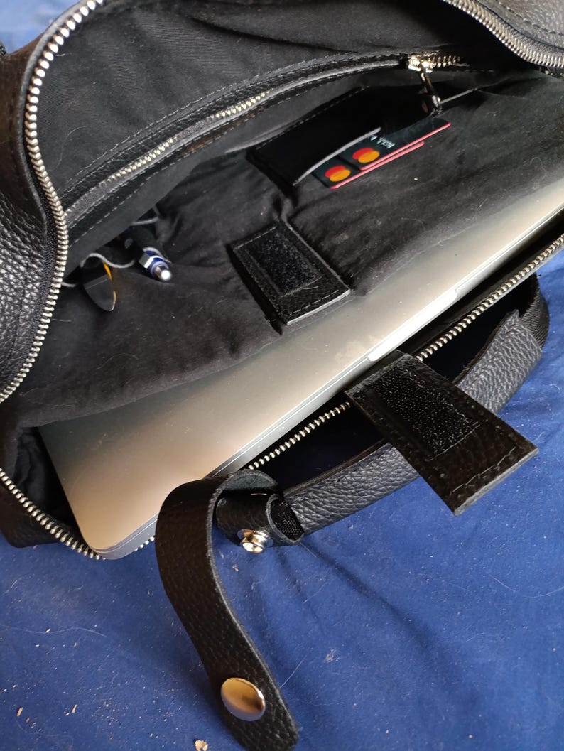 Puede incluir: Bolso para port&aacute;til de cuero negro con cremallera plateada y asa. El bolso est&aacute; abierto, revelando un port&aacute;til en el interior, junto con un peque&ntilde;o bolsillo que contiene una tarjeta de cr&eacute;dito. El interior es negro.