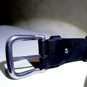 Op de afbeelding: Close-up van een zwarte leren riem met een zilverkleurige metalen gesp. De gesp is rechthoekig met een enkele metalen staaf. Het leer is glad en lijkt van hoge kwaliteit te zijn. De riem is ontworpen voor dagelijks gebruik.