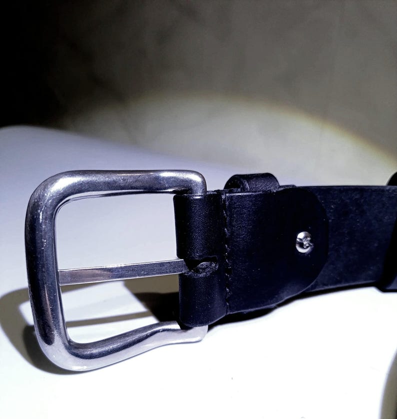 Op de afbeelding: Close-up van een zwarte leren riem met een zilverkleurige metalen gesp. De gesp is rechthoekig met een enkele metalen staaf. Het leer is glad en lijkt van hoge kwaliteit te zijn. De riem is ontworpen voor dagelijks gebruik.