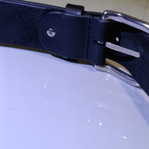 Op de afbeelding: Close-up van een zwarte leren riem met een zilverkleurige gesp. De riem heeft een eenvoudig ontwerp met een enkele lus en een metalen gesp. Het leer lijkt glad, geschikt voor dagelijks gebruik. De riem is ongeveer 3-4 cm breed.