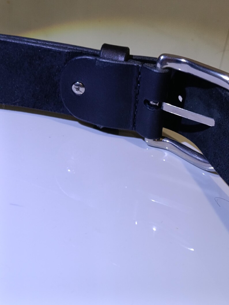 Op de afbeelding: Close-up van een zwarte leren riem met een zilverkleurige gesp. De riem heeft een eenvoudig ontwerp met een enkele lus en een metalen gesp. Het leer lijkt glad, geschikt voor dagelijks gebruik. De riem is ongeveer 3-4 cm breed.