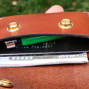 Puede incluir: Una cartera de cuero marr&oacute;n con dos botones de presi&oacute;n de metal dorado en la solapa. Dentro de la cartera, un encendedor verde brillante est&aacute; guardado junto a una tarjeta de cr&eacute;dito negra y un billete de cien d&oacute;lares doblado en el compartimento.