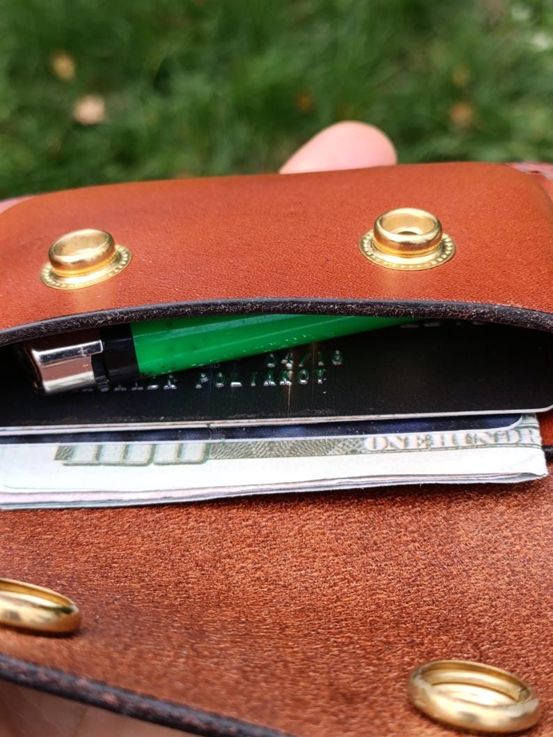 Puede incluir: Una cartera de cuero marr&oacute;n con dos botones de presi&oacute;n de metal dorado en la solapa. Dentro de la cartera, un encendedor verde brillante est&aacute; guardado junto a una tarjeta de cr&eacute;dito negra y un billete de cien d&oacute;lares doblado en el compartimento.