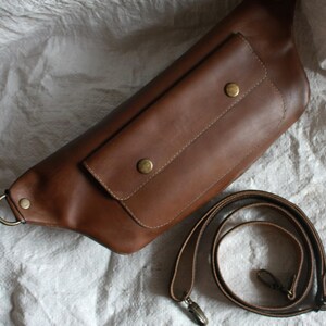 May include: A brown leather fanny pack with a large front pocket and a brown leather adjustable strap.