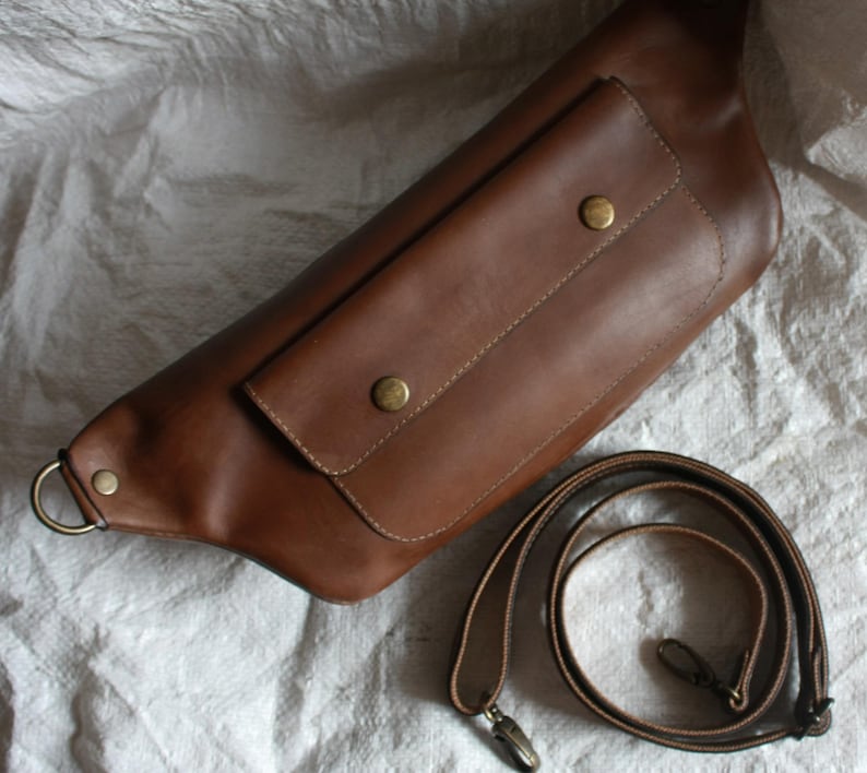 May include: A brown leather fanny pack with a large front pocket and a brown leather adjustable strap.