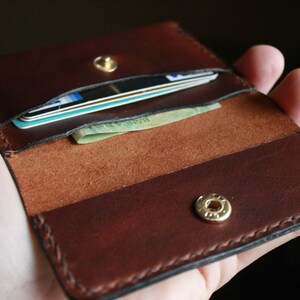 May include: Open brown leather wallet with cards and cash visible. The wallet has a snap closure and is held open, showcasing the interior compartments. The leather has a rich, dark brown color with visible stitching.