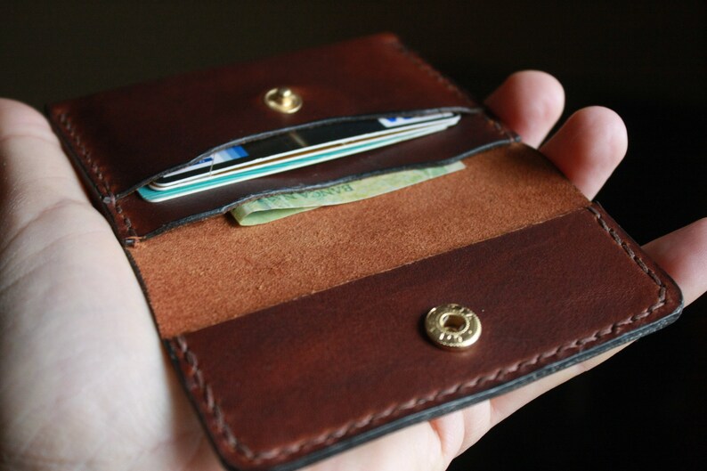 May include: Open brown leather wallet with cards and cash visible. The wallet has a snap closure and is held open, showcasing the interior compartments. The leather has a rich, dark brown color with visible stitching.