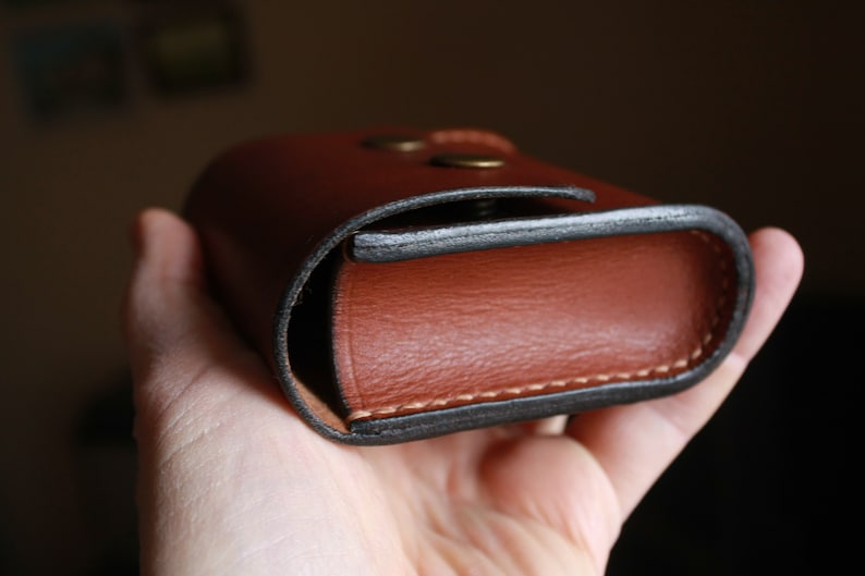 May include: A brown leather case with a black stitched edge and two brass rivets. The case is closed and held in a hand.