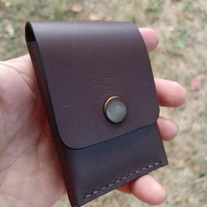May include: A dark brown leather card holder featuring a smooth flap closure secured by a single circular antique brass-toned metal snap button. The rectangular accessory is constructed with visible stitching along the bottom edge and sides.