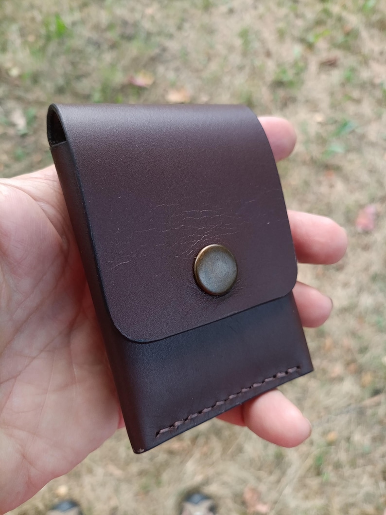 May include: A dark brown leather card holder featuring a smooth flap closure secured by a single circular antique brass-toned metal snap button. The rectangular accessory is constructed with visible stitching along the bottom edge and sides.