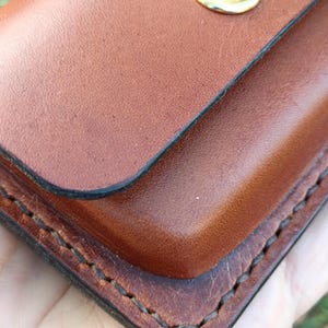 May include: A brown leather wallet featuring a smooth finish, dark burnished edges, and visible stitching along the perimeter. The wallet is secured with a single, polished gold-tone metal snap button closure centred on the top flap.