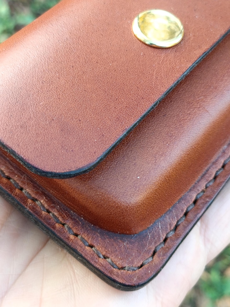 May include: A brown leather wallet featuring a smooth finish, dark burnished edges, and visible stitching along the perimeter. The wallet is secured with a single, polished gold-tone metal snap button closure centred on the top flap.