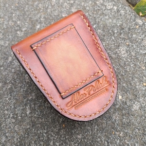 K&ouml;nnte beinhalten: Ein brauner Lederg&uuml;rtelclip mit einem rechteckigen R&uuml;ckenteil und einer gebogenen Vorderseite. Der Clip ist mit hellbraunem Faden gen&auml;ht. Der Text "Alex Polak Handmade Leather Goods" ist auf der Vorderseite gepr&auml;gt.