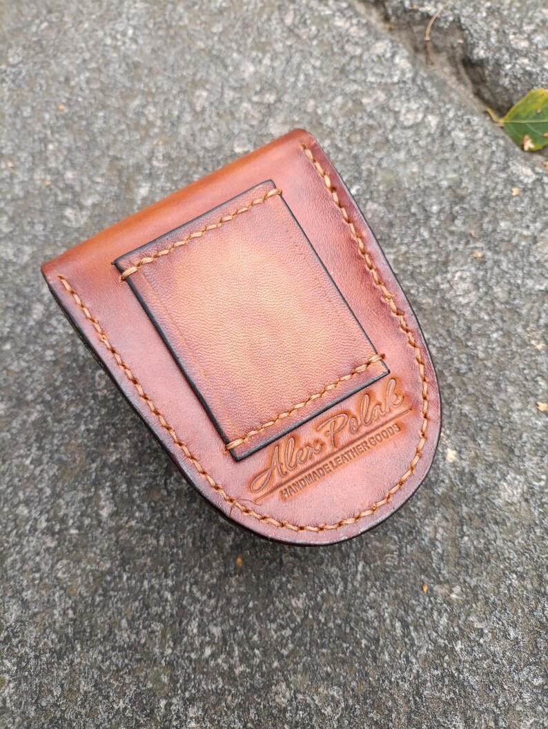 K&ouml;nnte beinhalten: Ein brauner Lederg&uuml;rtelclip mit einem rechteckigen R&uuml;ckenteil und einer gebogenen Vorderseite. Der Clip ist mit hellbraunem Faden gen&auml;ht. Der Text "Alex Polak Handmade Leather Goods" ist auf der Vorderseite gepr&auml;gt.