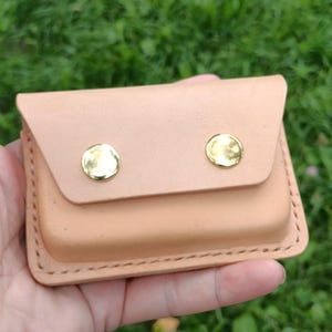 May include: A rectangular light tan leather card holder featuring a flap closure secured by two polished gold-tone metal snaps. The edges are stitched with thick thread, and the leather has a smooth, natural finish with visible grain details.
