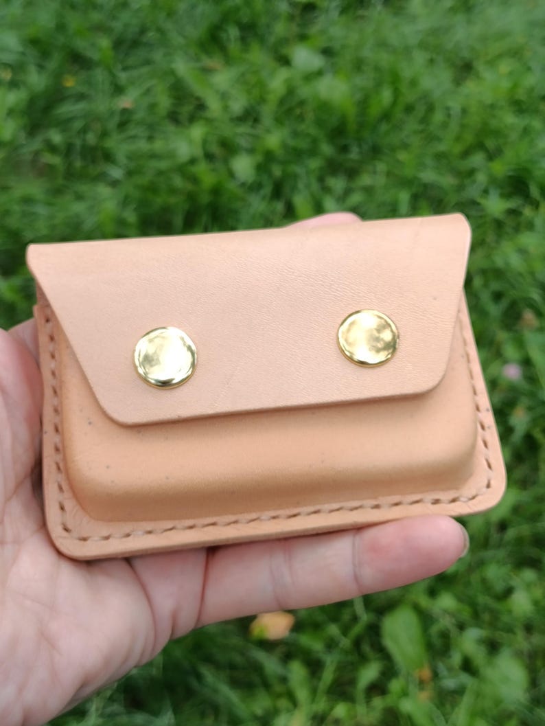 May include: A rectangular light tan leather card holder featuring a flap closure secured by two polished gold-tone metal snaps. The edges are stitched with thick thread, and the leather has a smooth, natural finish with visible grain details.