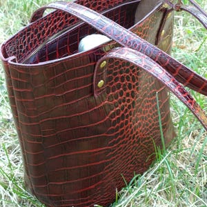 May include: A deep red tote bag with a textured crocodile skin pattern. The bag features two thin shoulder straps attached with gold-toned metal rivets, a zippered top closure, and a structured rectangular shape with a glossy finish.
