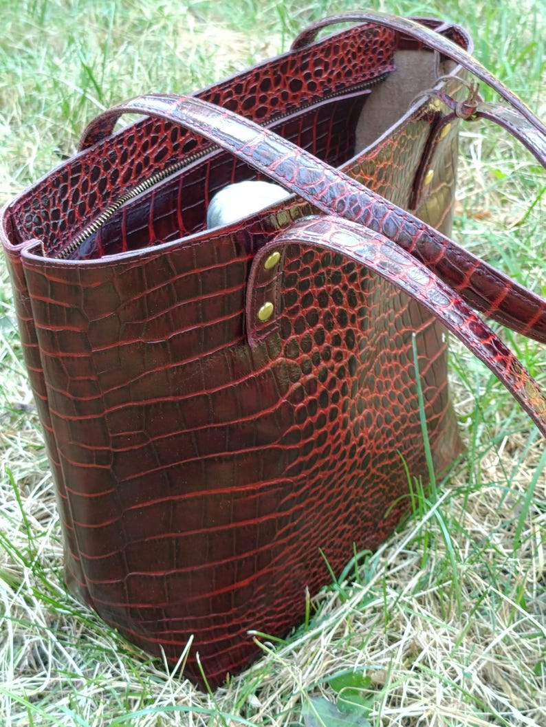 May include: A deep red tote bag with a textured crocodile skin pattern. The bag features two thin shoulder straps attached with gold-toned metal rivets, a zippered top closure, and a structured rectangular shape with a glossy finish.