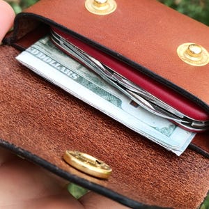 May include: A brown leather pouch with two gold-colored metal snap buttons on the flap. Inside the pouch, a red multi-tool and a folded United States one hundred dollar bill are visible. The interior of the leather shows a natural, textured grain.