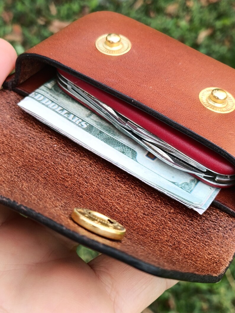 May include: A brown leather pouch with two gold-colored metal snap buttons on the flap. Inside the pouch, a red multi-tool and a folded United States one hundred dollar bill are visible. The interior of the leather shows a natural, textured grain.