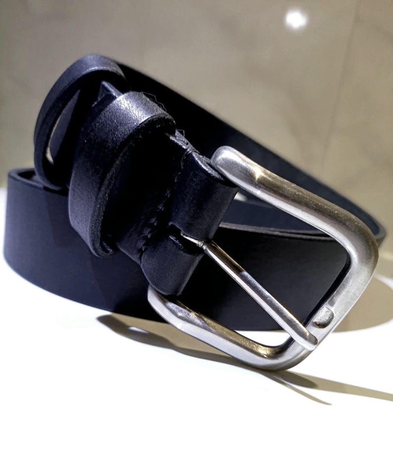 Op de afbeelding: Een zwarte leren riem met een zilverkleurige gesp. De riem is gemaakt van glad, donker leer en heeft een klassieke rechthoekige gesp. De riem is ontworpen voor dagelijks gebruik en is een veelzijdig accessoire.