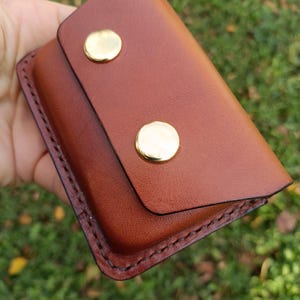 May include: A rectangular brown leather wallet featuring a flap closure secured by two polished gold-tone metal snaps. The edges of the wallet are finished with dark brown stitching, creating a defined border around the smooth leather surface.
