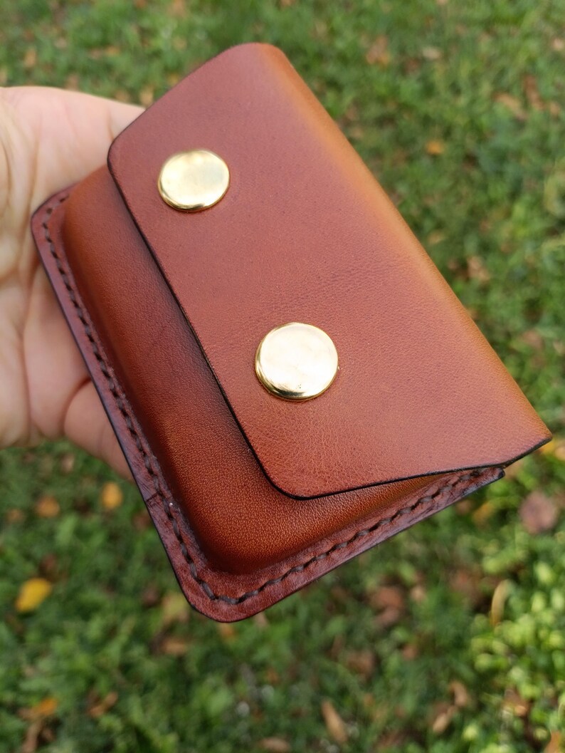 May include: A rectangular brown leather wallet featuring a flap closure secured by two polished gold-tone metal snaps. The edges of the wallet are finished with dark brown stitching, creating a defined border around the smooth leather surface.