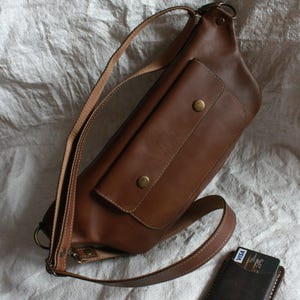 May include: A brown leather crossbody bag with a large front pocket and a smaller pocket on the flap. The bag has a long adjustable strap and a metal buckle.