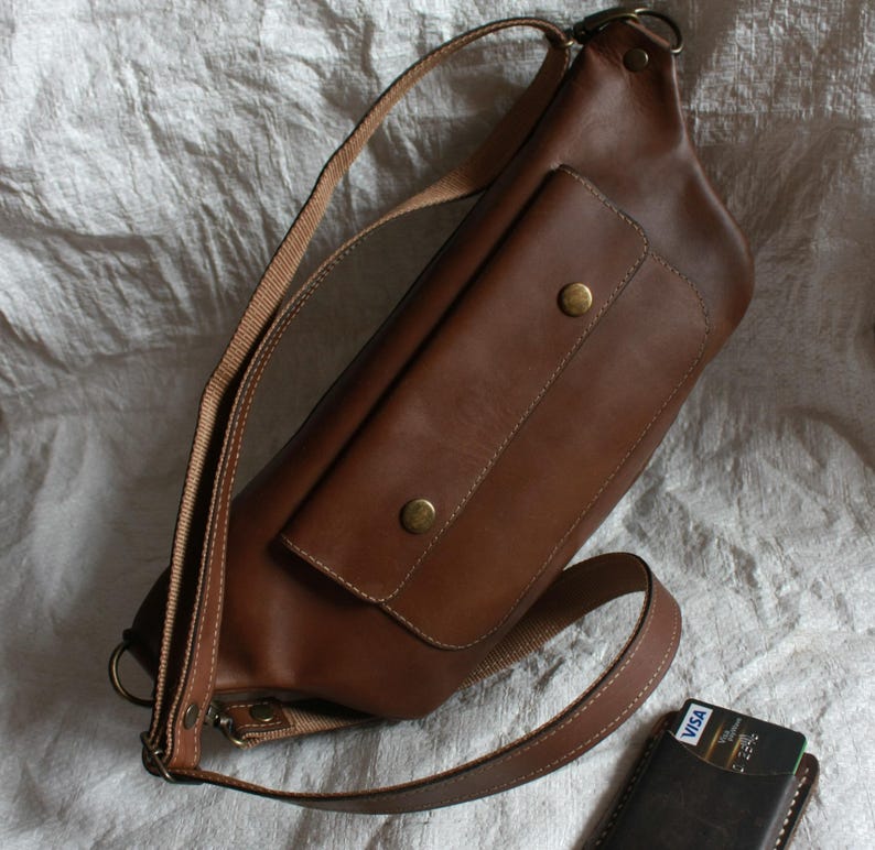 May include: A brown leather crossbody bag with a large front pocket and a smaller pocket on the flap. The bag has a long adjustable strap and a metal buckle.