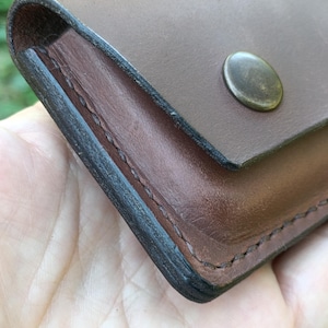 Puede incluir: Una cartera de cuero marr&oacute;n con una superficie lisa, bordes oscuros bru&ntilde;idos y un cierre de bot&oacute;n a presi&oacute;n met&aacute;lico. La cartera est&aacute; construida con costuras visibles a lo largo del lateral, mostrando un dise&ntilde;o rectangular simple.