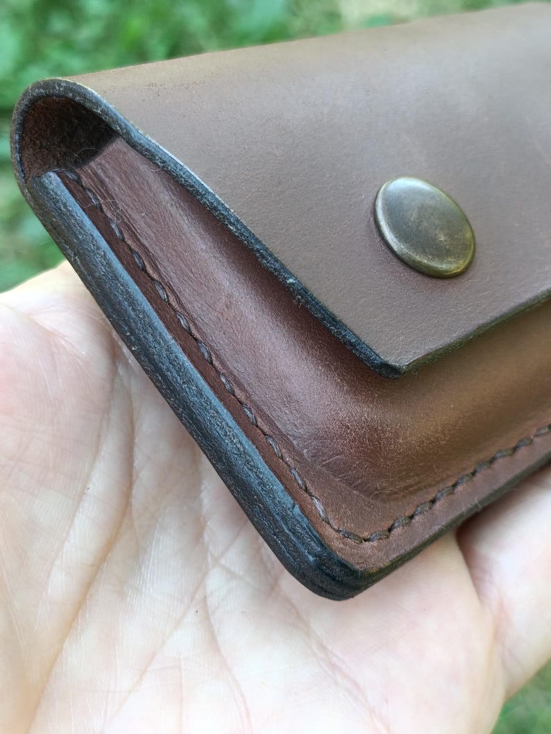 Puede incluir: Una cartera de cuero marr&oacute;n con una superficie lisa, bordes oscuros bru&ntilde;idos y un cierre de bot&oacute;n a presi&oacute;n met&aacute;lico. La cartera est&aacute; construida con costuras visibles a lo largo del lateral, mostrando un dise&ntilde;o rectangular simple.