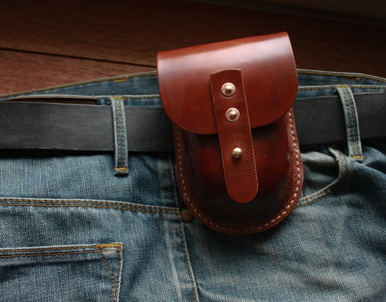 May include: A brown leather pouch with a curved top, secured with two rivets and a strap, attached to a belt over blue denim jeans. The pouch is stitched with white thread.