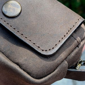 Peut inclure: Une pochette marron fonc&eacute; avec une surface mate et textur&eacute;e, dot&eacute;e d'un rabat ferm&eacute; par un bouton en laiton de style antique. Les bords du rabat pr&eacute;sentent des coutures marron fonc&eacute; visibles, et une tirette de fermeture &eacute;clair est visible.