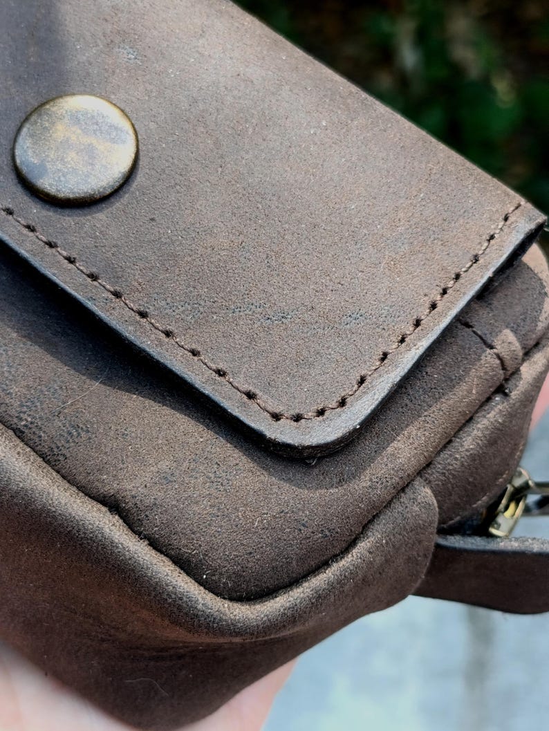 Peut inclure: Une pochette marron fonc&eacute; avec une surface mate et textur&eacute;e, dot&eacute;e d'un rabat ferm&eacute; par un bouton en laiton de style antique. Les bords du rabat pr&eacute;sentent des coutures marron fonc&eacute; visibles, et une tirette de fermeture &eacute;clair est visible.