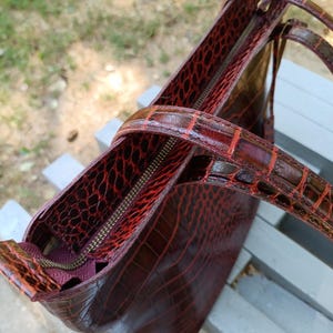 May include: A deep burgundy tote bag featuring a glossy, embossed crocodile skin texture. The bag includes two matching shoulder straps, a top zipper closure with a metallic pull, and a dark interior lining visible through the open top section.