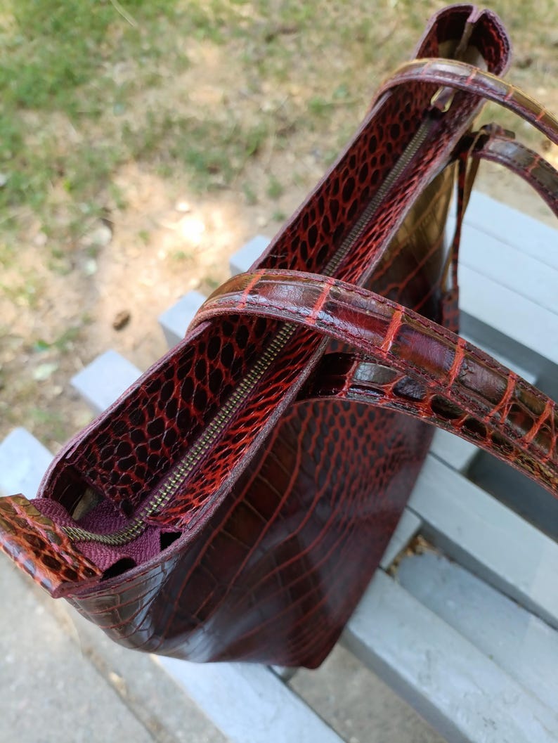 May include: A deep burgundy tote bag featuring a glossy, embossed crocodile skin texture. The bag includes two matching shoulder straps, a top zipper closure with a metallic pull, and a dark interior lining visible through the open top section.