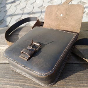 Pu&ograve; includere: Una borsa a tracolla in pelle marrone con chiusura a lembo e fibbia. La borsa &egrave; aperta e l'interno &egrave; foderato con un tessuto marrone chiaro.