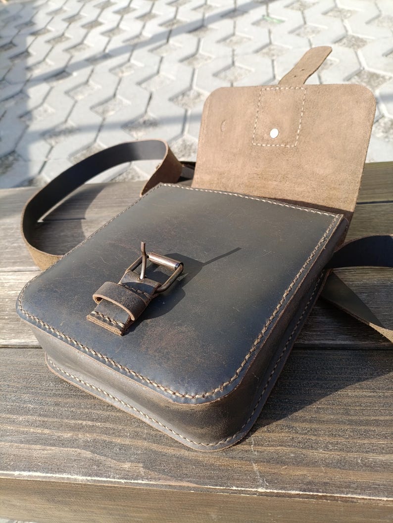 Pu&ograve; includere: Una borsa a tracolla in pelle marrone con chiusura a lembo e fibbia. La borsa &egrave; aperta e l'interno &egrave; foderato con un tessuto marrone chiaro.