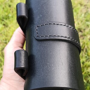 May include: A black leather holster with a cylindrical shape, featuring a stitched strap and two loops. The holster is designed to hold an item and is held in a hand against a blurred green background.