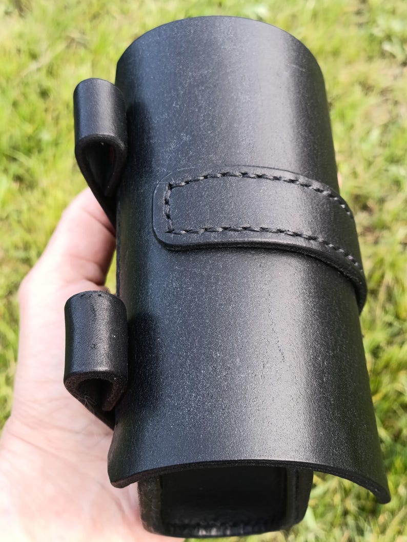 May include: A black leather holster with a cylindrical shape, featuring a stitched strap and two loops. The holster is designed to hold an item and is held in a hand against a blurred green background.