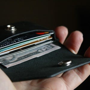 Puede incluir: Una cartera de cuero negro con cierre a presi&oacute;n, que contiene varias tarjetas de cr&eacute;dito y efectivo. La cartera est&aacute; abierta y se sostiene en una mano.