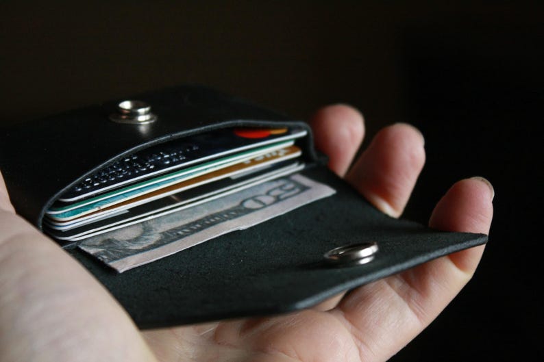Puede incluir: Una cartera de cuero negro con cierre a presi&oacute;n, que contiene varias tarjetas de cr&eacute;dito y efectivo. La cartera est&aacute; abierta y se sostiene en una mano.