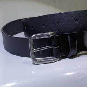 Op de afbeelding: Een zwarte leren riem met een zilverkleurige gesp. De riem heeft een klassiek ontwerp met een rechthoekige gesp en meerdere gaten voor verstelling. Het leer ziet er glad en duurzaam uit, geschikt voor dagelijks gebruik.