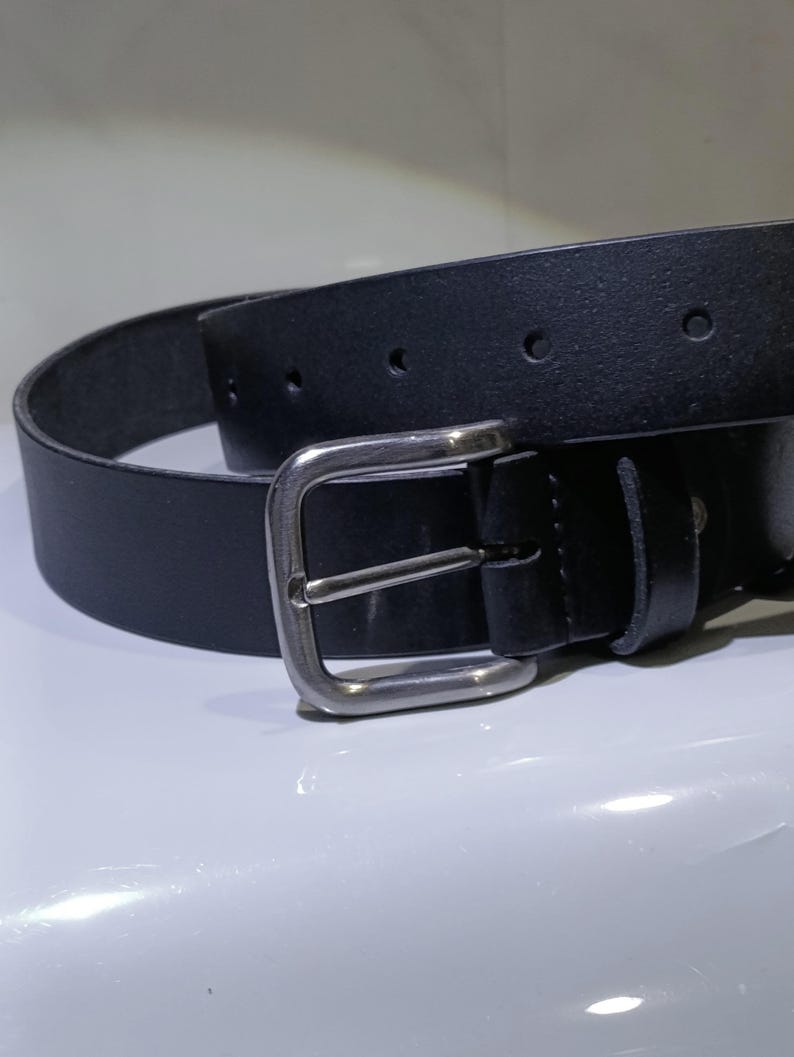 Op de afbeelding: Een zwarte leren riem met een zilverkleurige gesp. De riem heeft een klassiek ontwerp met een rechthoekige gesp en meerdere gaten voor verstelling. Het leer ziet er glad en duurzaam uit, geschikt voor dagelijks gebruik.