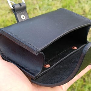 May include: Black leather pouch with a silver buckle closure. The open pouch reveals a small interior compartment with copper rivets. The pouch is held against a background of green grass.