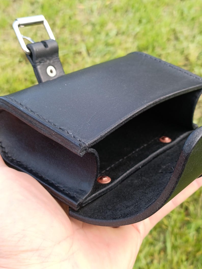 May include: Black leather pouch with a silver buckle closure. The open pouch reveals a small interior compartment with copper rivets. The pouch is held against a background of green grass.