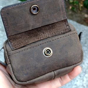 Peut inclure: Une petite pochette rectangulaire en cuir marron fonc&eacute; dot&eacute;e d'un rabat avec une fermeture &agrave; bouton-pression m&eacute;tallique. Le cuir pr&eacute;sente une finition mate et textur&eacute;e avec des coutures visibles sur les bords et une attache centrale.