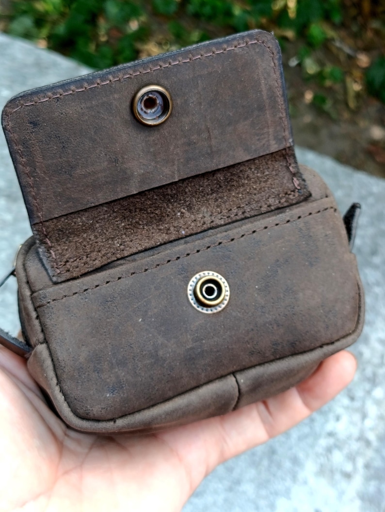 Peut inclure: Une petite pochette rectangulaire en cuir marron fonc&eacute; dot&eacute;e d'un rabat avec une fermeture &agrave; bouton-pression m&eacute;tallique. Le cuir pr&eacute;sente une finition mate et textur&eacute;e avec des coutures visibles sur les bords et une attache centrale.