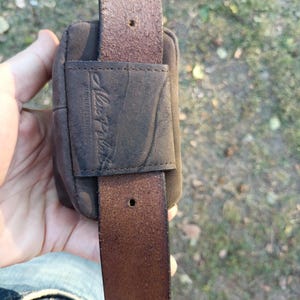 Peut inclure: Une pochette de ceinture en cuir marron fonc&eacute; dot&eacute;e d'une boucle rectangulaire en cuir sur le devant. La boucle est gaufr&eacute;e avec le texte Alex Plus et comprend une sangle en cuir verticale qui la traverse. Le cuir a une finition mate et textur&eacute;e.